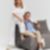 Um homem de roupa elegante sentado num sofá cinza de maneira despojada. Ao lado dele está uma mulher loira encostada no encosto do sofá. Os dois estão num local com fundo totalmente branco